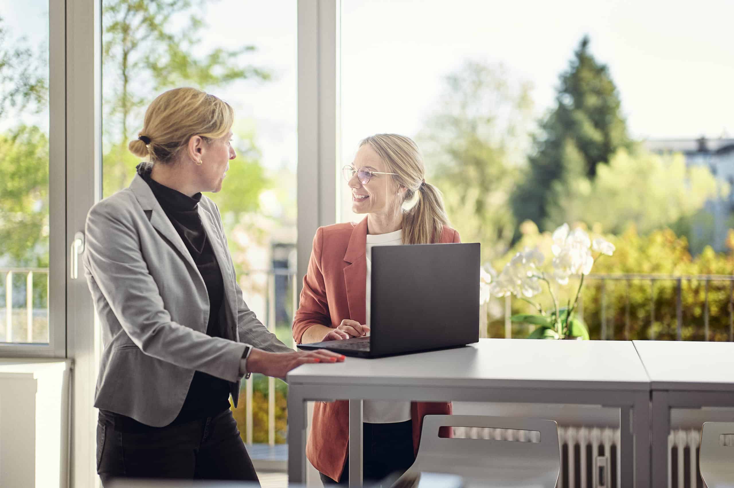 Celine Spitz and Magdalena Sabaß are standing in a lounge at a bar table with a laptop on it, chatting with each other.