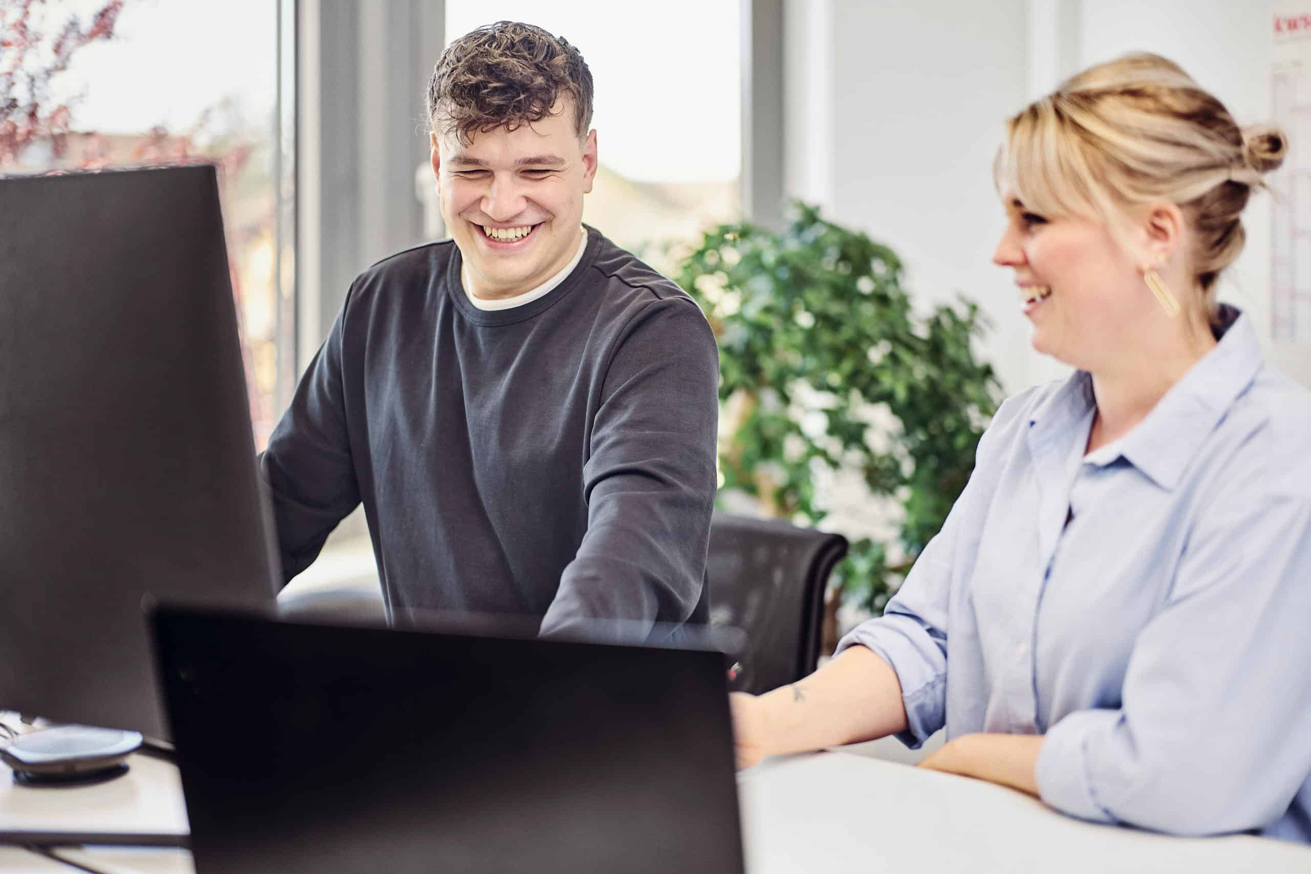 Julius Binninger and Anna Wißler are standing at the office desk, smiling as they look at the screens.