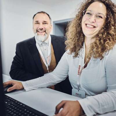 A woman in a blue shirt and a man in a black suit are sitting at a computer and laughing at the camera.