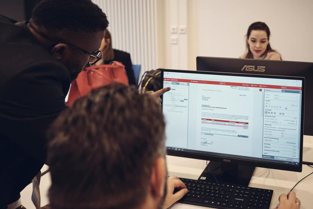 One man sits at the computer using software, while another man in a suit points at the screen and explains what is happening.