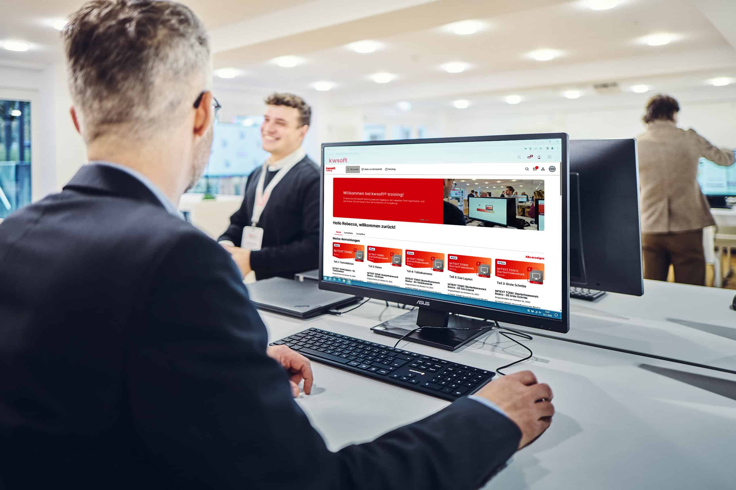 A man sits in front of a screen running kwsoft training and looks at a man standing in the background.