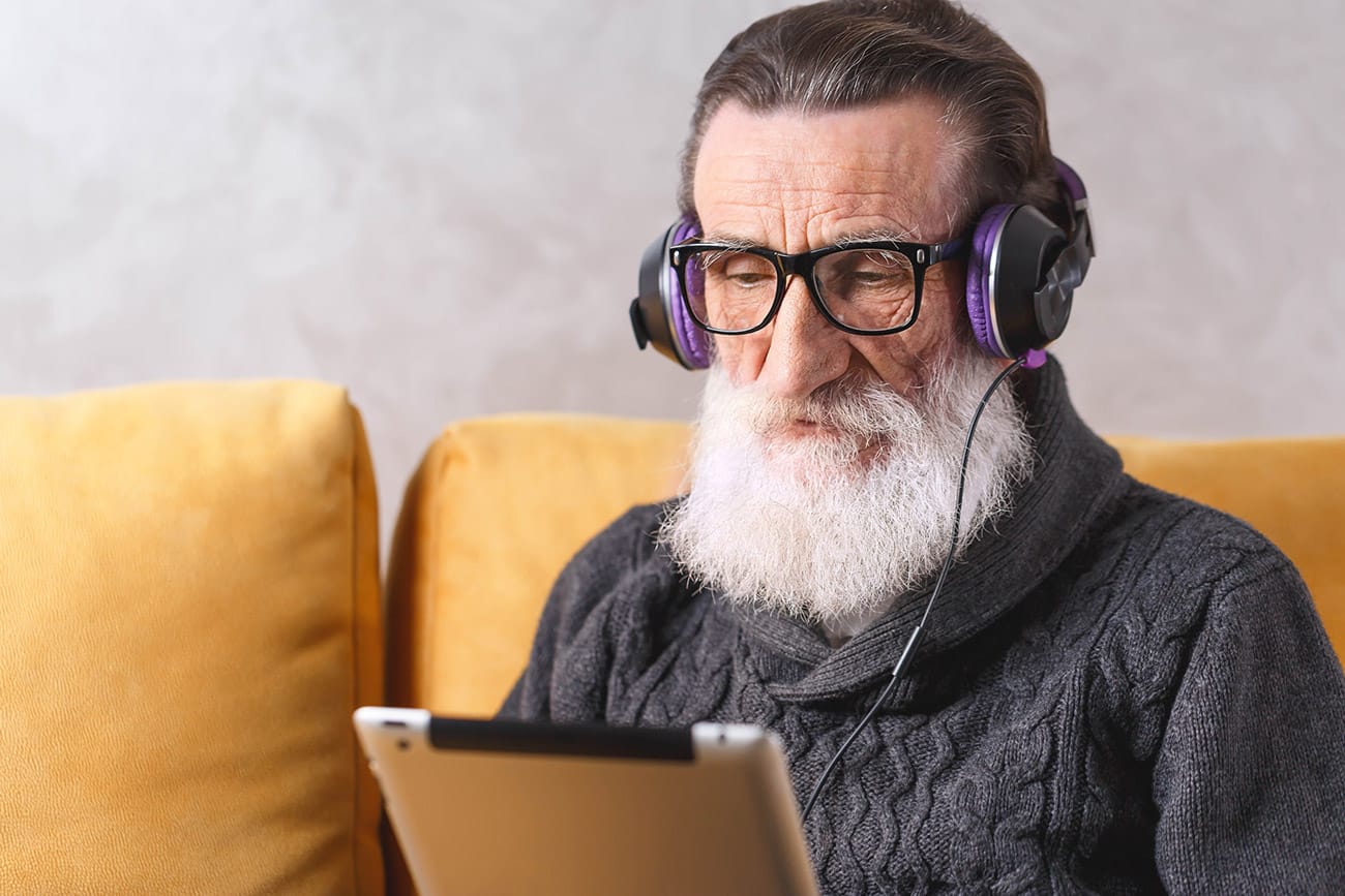 An older man with glasses is sitting on a couch wearing headphones connected to the tablet he is holding in his hand.